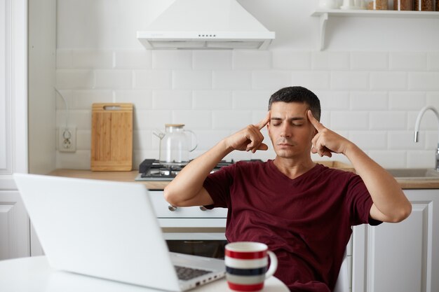 Toma interior de un hombre agotado posando en la cocina mientras está sentado a la mesa frente a la computadora portátil, se siente cansado, mantiene los ojos cerrados, se masajea las sienes con los dedos, sufre de dolor de cabeza.