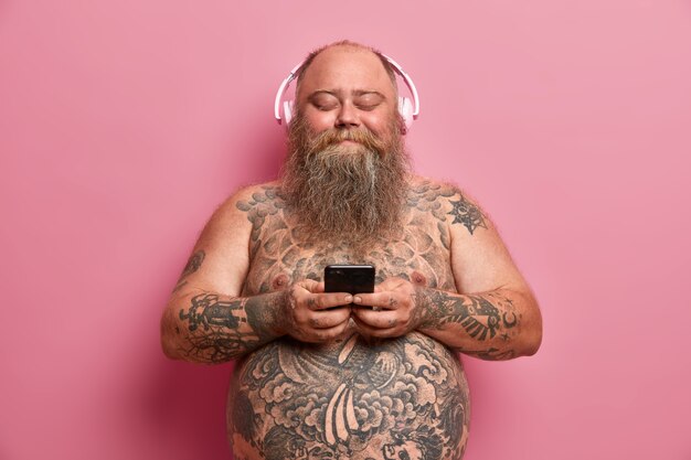 Toma interior de un hombre gordo adulto satisfecho que sostiene el teléfono móvil, usa la aplicación de música, usa auriculares estéreo en los oídos, escucha canciones, está desnudo, tiene el cuerpo tatuado, aislado en una pared rosa. Exceso de peso