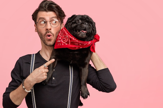 Toma interior de un hombre guapo asombrado con expresión estupefacta, vestido con ropa elegante, pasa tiempo libre con su perro favorito, apunta al espacio libre contra la pared rosa. Hombre con mascota