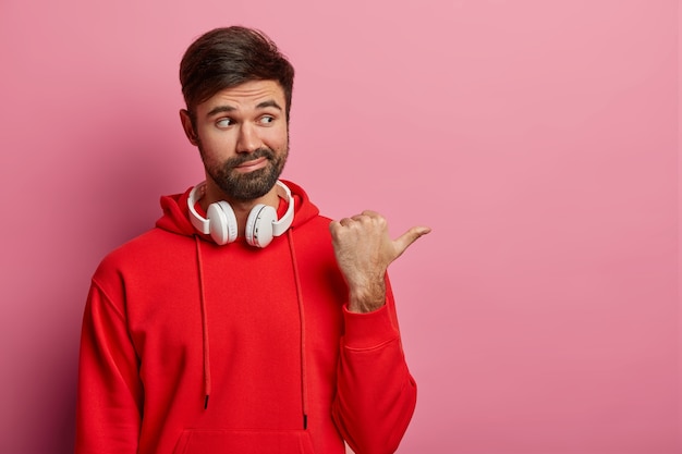 La toma interior de un hombre indeciso sin afeitar indica a un lado, tiene una mirada dudosa a un lado, pregunta si debe ir allí, usa audífonos alrededor del cuello, vestido con una sudadera con capucha roja, posa contra la pared rosa pastel