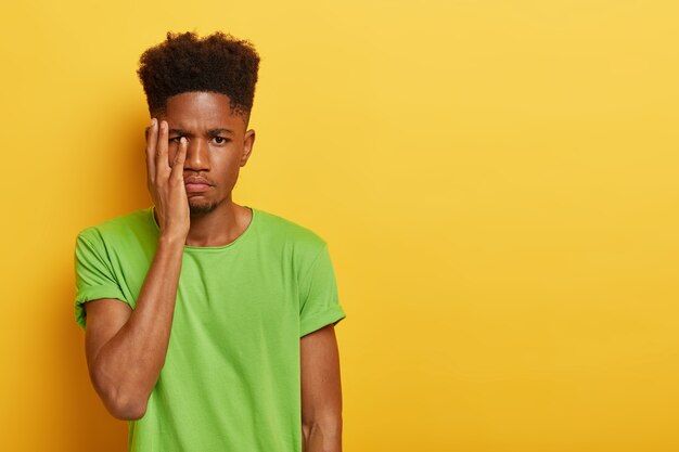 Toma interior de un hombre de piel oscura molesto con corte de pelo afro, se cubre la cara con la palma, tiene expresión facial cansada, trabaja largas horas preparándose para el examen, tiene fecha límite, usa camiseta verde