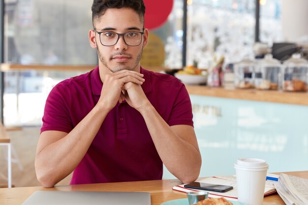 Toma interior de un hombre de raza mixta que mantiene las manos debajo de la barbilla, se ve con expresión facial segura de sí mismo, usa gafas ópticas, se sienta a la mesa en una acogedora cafetería, toma un café después del trabajo remoto en la computadora portátil