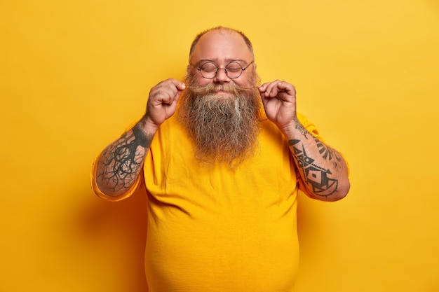 Foto gratuita toma interior de hombre regordete complacido gira bigote, se jacta de barba espesa, se para con los ojos cerrados, sonríe agradablemente, tiene brazos tatuados vestidos con ropa amarilla usa gafas pequeñas redondas posa interior