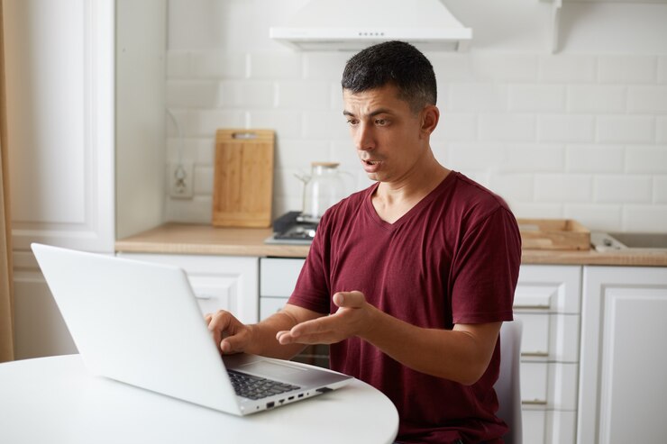 Foto gratuito toma interior de un joven adulto independiente sentado en la cocina en la mesa blanca, trabajando en línea en la computadora, teniendo problemas con el trabajo, mirando la pantalla de la pc con expresión de desconcierto.