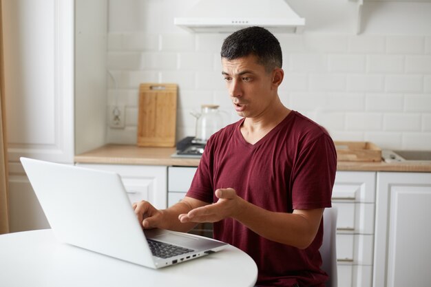 Toma interior de un joven adulto independiente sentado en la cocina en la mesa blanca, trabajando en línea en la computadora, teniendo problemas con el trabajo, mirando la pantalla de la PC con expresión de desconcierto.