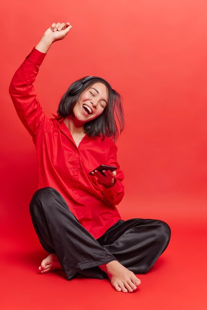 Toma interior de una joven asiática despreocupada sentada con las piernas cruzadas en el suelo, mantiene el brazo levantado, sostiene el teléfono móvil, escucha música a través de los auriculares, usa camisa y pantalones negros aislados sobre un fondo rojo