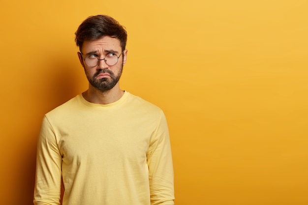 Toma interior de un joven barbudo molesto que se ve con expresión miserable a un lado, piensa en algo, tiene una mirada triste e infeliz, usa lentes ópticos y un jersey casual, posa en el interior sobre una pared amarilla