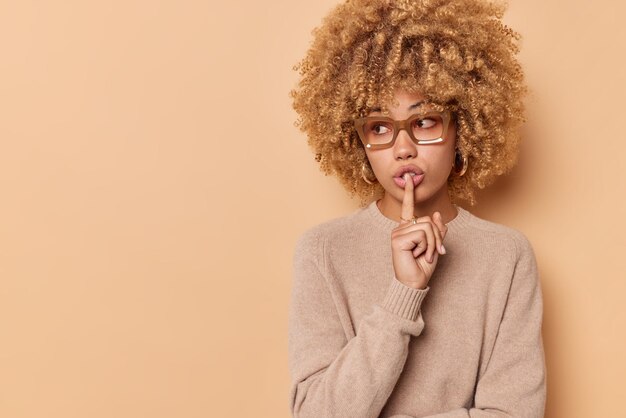 La toma interior de una joven de cabello rizado hace un gesto de silencio que presiona el dedo índice contra los labios y mira misteriosamente hacia otro lado vestida casualmente aislada sobre un espacio de copia en blanco de fondo beige. Es secreto