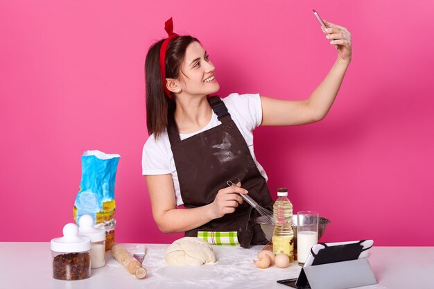 Toma interior de una joven carismática smilling de pie haciendo selfie en la cocina mientras cocina un plato nuevo y delicioso, publicando fotos y videos en sitios de redes sociales. Concepto de cocción y horneado.