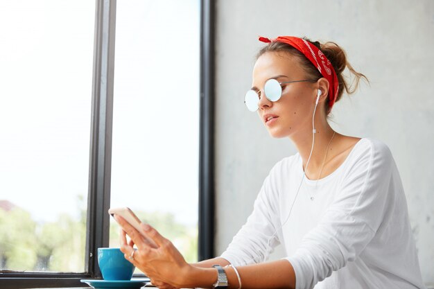 Toma interior de una joven linda que lleva una diadema roja y gafas de sol, escucha la composición favorita de la lista de reproducción a través del teléfono móvil, conectado a internet inalámbrico y auriculares en una acogedora cafetería