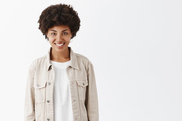 Toma interior de una linda chica de piel oscura y peinado afro con una moderna chaqueta beige sobre una camiseta, sonriendo ampliamente y mirando con expresión segura y despreocupada, sintiéndose feliz y relajada