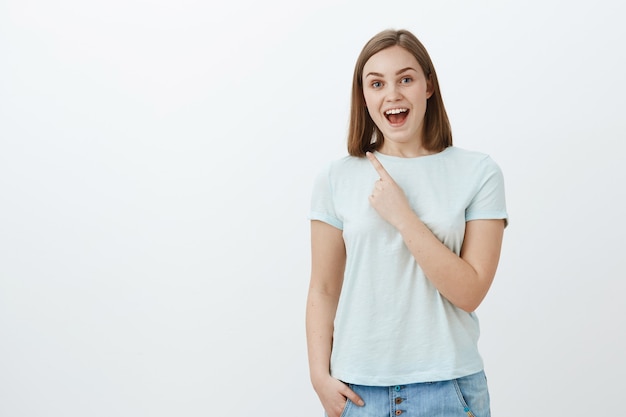 Toma interior de una linda mujer asombrada, energizada y optimista en una camiseta de moda sonriendo alegremente apuntando a la esquina superior izquierda con el dedo índice cerca del pecho disfrutando de pasar tiempo divertido y divertido