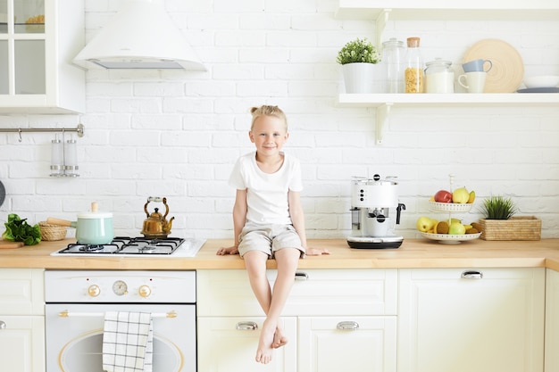 Toma interior de un lindo niño guapo con un moño de pelo rubio sentado en el mostrador de la elegante cocina, colgando de sus pies descalzos, esperando que la madre prepare el desayuno en la mañana antes de la escuela