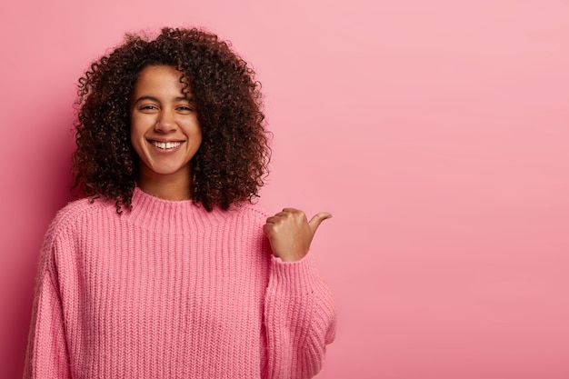 La toma interior de una mujer afro satisfecha tiene el cabello oscuro y rizado, señala a un lado con el pulgar, muestra un bonito espacio para copiar su publicidad, tiene una expresión amistosa, viste un suéter rosa de gran tamaño.