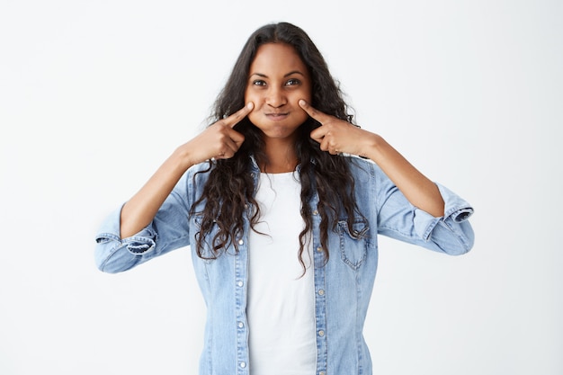 Toma interior de mujer afroamericana bonita morena haciendo una mueca soplando sus mejillas con expresión complacida. Hermosa mujer joven con piel oscura vestida con ropa casual burlándose.