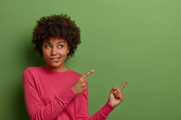 Toma interior de una mujer afroamericana curiosa que se muerde los labios, ve algo bueno, indica los dedos en un espacio en blanco en la pared verde, muestra el lugar de su producto, banner de la empresa o promoción