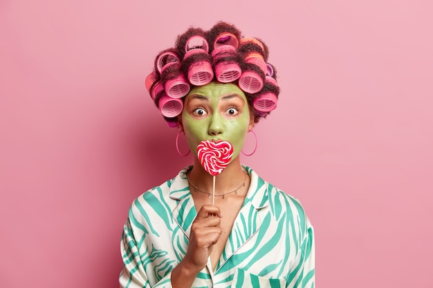La toma interior de una mujer afroamericana sorprendida aplica rulos para hacer que el peinado perfecto cubra la boca con una paleta que hace una máscara nutritiva verde para reducir las líneas finas aisladas sobre la pared rosa.