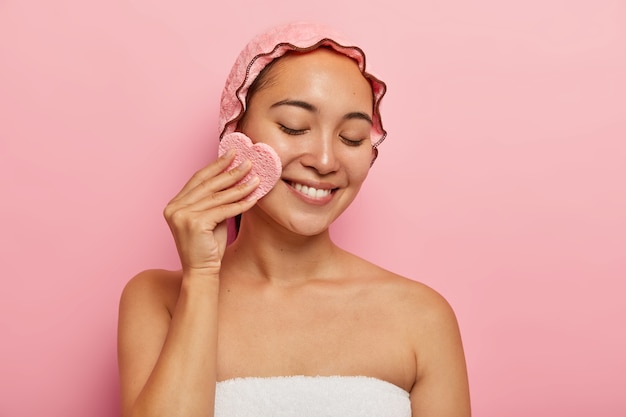 Toma interior de una mujer asiática bastante joven que usa una esponja cosmética para desmaquillarse, tiene una piel grasa problemática, enfocada hacia abajo con una sonrisa tierna, envuelta en una toalla aislada en una pared rosa. concepto de belleza