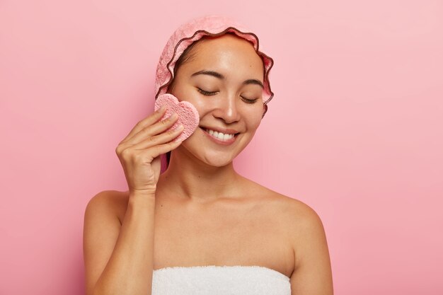 Toma interior de una mujer asiática bastante joven que usa una esponja cosmética para desmaquillarse, tiene una piel grasa problemática, enfocada hacia abajo con una sonrisa tierna, envuelta en una toalla aislada en una pared rosa. Concepto de belleza