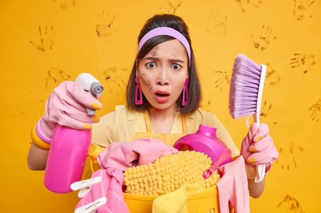 Toma interior de mujer asiática disgustada ocupada haciendo tareas domésticas estacionales usa detergente para lavar ventanas limpia muebles con cepillo lava la ropa tiene la cara sucia usa guantes de goma diadema