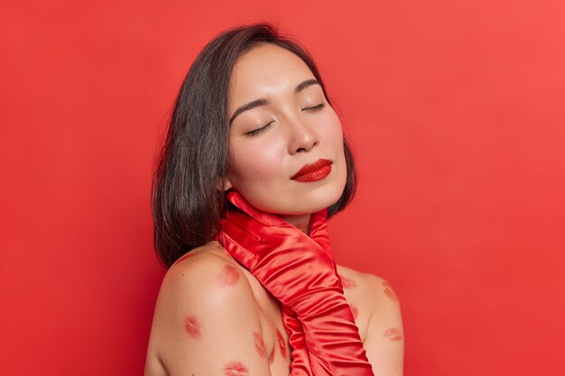 Toma interior de mujer asiática con maquillaje natural, lápiz labial rojo mantiene la mano en el cuello sin camisa tiene rastros de lápiz labial en el cuerpo aislado contra la pared vívida
