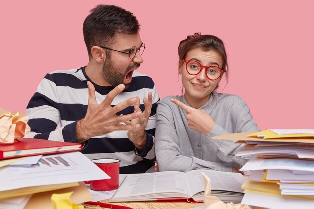 Toma interior de una mujer bonita que apunta a un novio enojado, lo culpa por el fracaso, estudian juntos, se preparan para la prueba final en la universidad, leen libros de texto, posan en el espacio de coworking