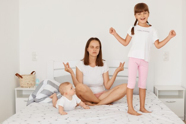 Toma interior de una mujer con cabello oscuro sentada en posición de loto y haciendo ejercicios de yoga mientras sus dos hijas, un bebé y una niña mayor juegan cerca de la madre.