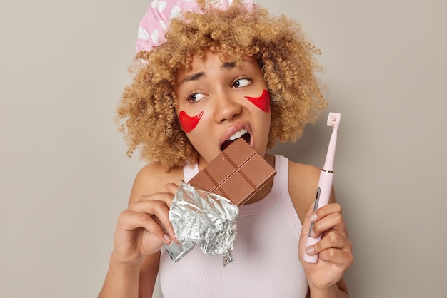 La toma interior de una mujer de cabello rizado muerde chocolate dulce dañino para los dientes sostiene un cepillo de dientes eléctrico se somete a procedimientos de belleza aplica parches rojos debajo de las poses de los ojos contra el fondo gris del estudio