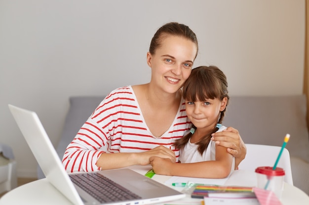 Toma interior de mujer feliz positiva con hija, sentada a la mesa con computadora portátil y libros, mujer abrazando a su hijo, gente mirando cámara de arte con expresión optimista.