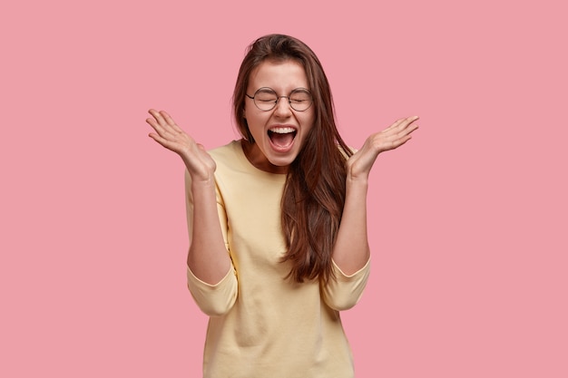 Toma interior de mujer joven feliz emocional cierra los ojos de placer, gestos, siendo muy emocional, vestida de manera informal, posa sobre la pared rosa. Concepto de personas, felicidad y positividad.