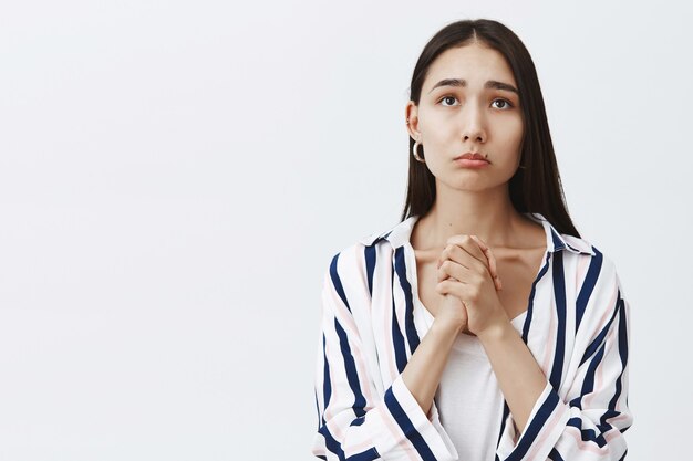 Toma interior de una mujer linda y sombría con una blusa a rayas sobre una camiseta, uniendo las manos y frunciendo los labios, frunciendo el ceño mientras mira hacia arriba con expresión esperanzada, esperando los resultados de la prueba sobre una pared gris