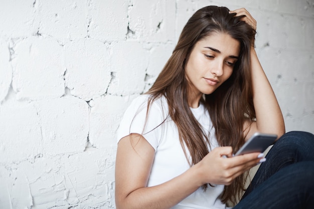 Toma interior de una mujer morena linda inteligente, apoyado en una pared de ladrillo mientras está sentado en el piso y navega por internet a través del teléfono inteligente.