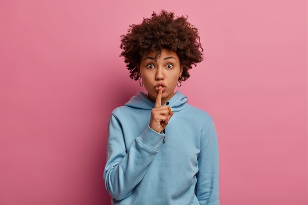 La toma interior de una mujer morena sorprendida tiene un plan secreto, hace un gesto de silencio, mira con expresión estupefacta, viste una sudadera con capucha azul, muestra un letrero de silencio o silencio, posa en el interior sobre una pared rosada.