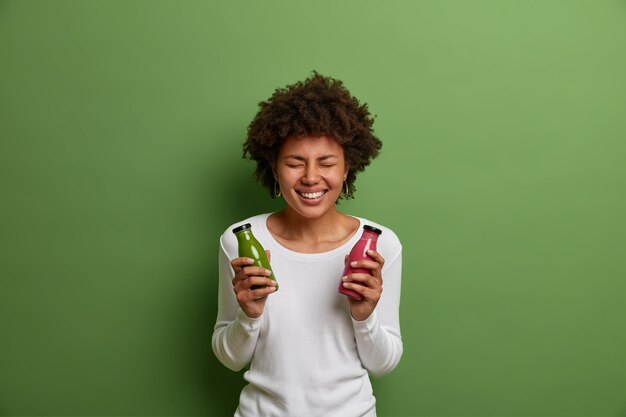 Toma interior de mujer sonriente muy feliz sostiene botellas de licuado de espinacas y fresa mezcladas, disfruta bebiendo bebidas de desintoxicación, posa con bebida fresca recién preparada, aislada sobre fondo verde