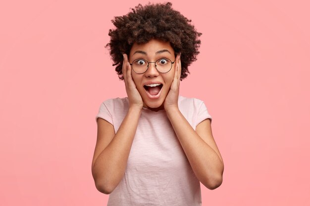 Toma interior de una mujer sorprendida y alegre con apariencia atractiva, cabello negro rizado y piel oscura, mira fijamente con ojos llenos de incredulidad, mantiene las manos en las mejillas, recibe algo inesperado y costoso