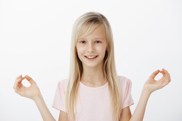 Toma interior de una niña adorable y curiosa con cabello rubio, extendiendo las manos en gestos zen y sonriendo con expresión satisfecha, meditando o practicando yoga, de pie sobre la pared gris tranquila