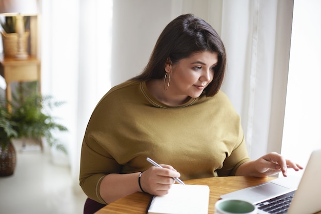 Toma interior de sobrepeso más tamaño hermosa joven morena con ropa elegante sentada en el escritorio con computadora portátil abierta, taza de café y anotando información en su diario, estudiando en línea