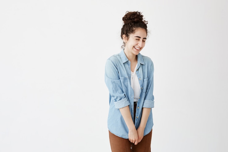 Foto gratuito toma interior de tímida chica linda estudiante con moño sonriendo alegremente, en camisa vaquera y pantalones marrones satisfechos con el presente. emociones positivas, sentimientos y expresiones faciales.