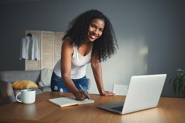 Toma en interiores de una atractiva joven positiva de raza mixta con cabello negro rizado sonriendo ampliamente, haciendo planes para el día, escribiendo en un cuaderno por la mañana antes de ir a trabajar