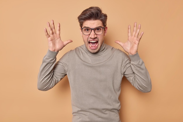 La toma en interiores de un chico moreno emocional mantiene las palmas levantadas, gritos indignados por estar molesto con alguien vestido con un jersey informal que se siente enojado aislado sobre un fondo beige. Concepto de emociones negativas