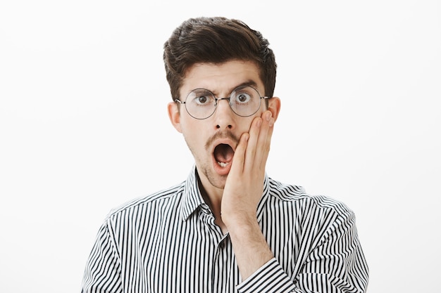 Foto gratuita toma en interiores de un compañero de trabajo masculino adulto emotivo conmocionado con gafas, dejando caer la mandíbula y sosteniendo la palma en la mejilla, sorprendido y aturdido con la historia veraz del empleado, de pie asombrado sobre una pared gris