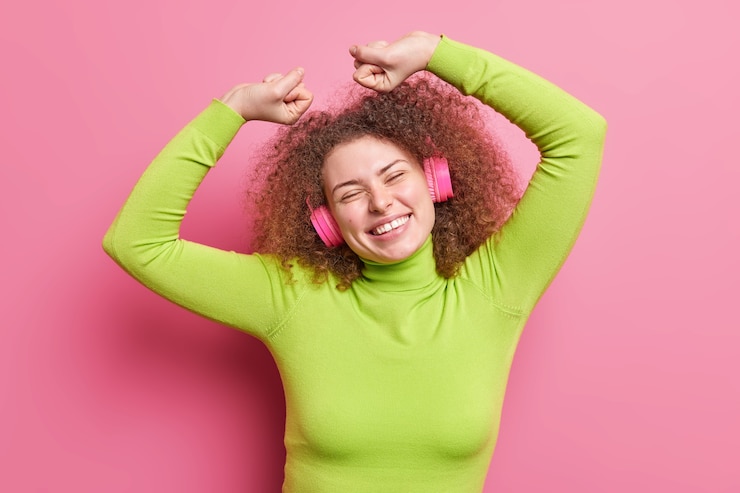 Foto gratuito toma en interiores de una despreocupada chica europea con cabello rizado levanta los brazos, aprieta los puños y celebra el éxito vestida con movimientos casuales de salto con ritmo de música, usa auriculares inalámbricos posa sobre una pared rosa
