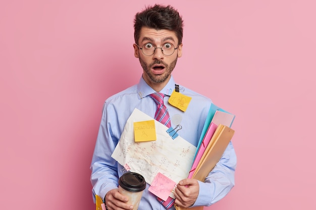 Toma en interiores de estudiantes masculinos calificados conmocionados que miran fijamente con los ojos abiertos y la mandíbula caída trabaja en el proyecto o se prepara para los exámenes escribe información en papeles para recordar bebidas café para llevar