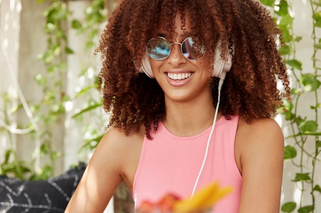 Toma en interiores de una hermosa mujer joven con peinado afro, usa gafas de sol de moda, escucha sus canciones favoritas solo con auriculares, tiene una sonrisa brillante. Mujer de piel oscura despreocupada se ríe alegremente