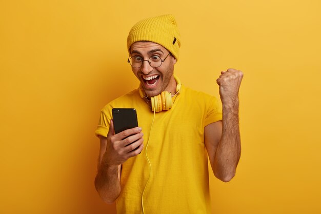 La toma en interiores de un hombre feliz muestra un golpe de puño con alegría, usa un teléfono celular, recibe noticias fantásticas, celebra su promoción, usa lentes transparentes