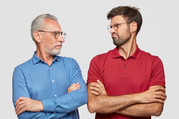 Toma en interiores de un joven disgustado que mira a su abuelo, mantiene las manos cruzadas, habla de la vida, se para uno al lado del otro, aislado sobre una pared blanca. Personas, concepto de comunicación