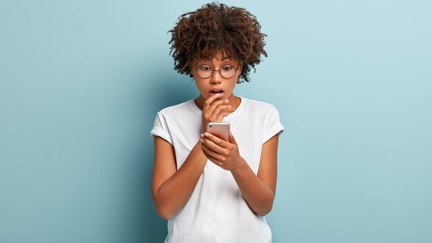 Toma en interiores de una mujer afroamericana aturdida enfocada en el teléfono celular, lee noticias terribles e impresionantes, usa una aplicación moderna, usa anteojos para corregir la visión aislada contra la pared azul tiene una llamada perdida