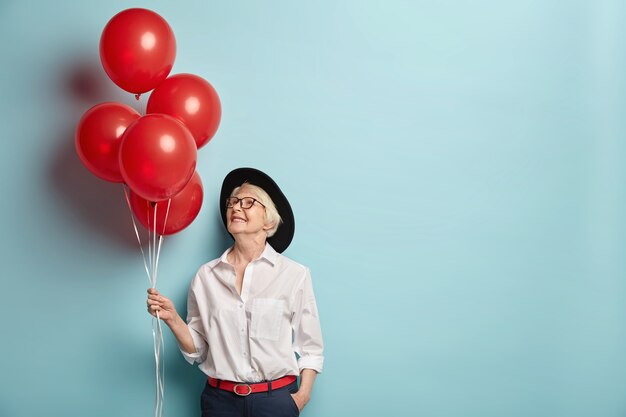 Toma en interiores de una mujer alegre de aspecto agradable de la vejez que sostiene globos de helio, viene en una fiesta para personas mayores para felicitar a su mejor amiga, enfocada arriba, usa ropa de moda