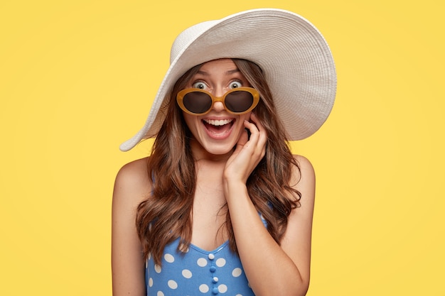 Toma en interiores de una mujer europea de aspecto agradable que tiene una sonrisa agradable, usa sombrero de verano, gafas de sol y vestido, se alegra de tener un viaje inolvidable, posa en una pared amarilla. Concepto de moda