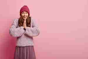 Foto gratuita la toma en interiores de una mujer feliz y complacida mantiene las palmas juntas, reza y espera algo mejor, tiene una expresión facial alegre, vestida con ropa cálida a la moda, se para sobre una pared rosada con espacio libre